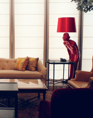 h. A red lamp casts a warm glow on a coffee table, and Roman shades filter sunlight through the window.