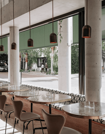 A stylish cafe interior with large windows featuring green motorized shades. The shades are partially lowered, creating a warm and inviting atmosphere with filtered sunlight streaming in.