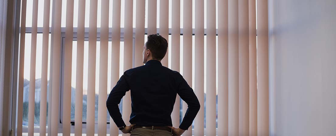 A man is standing in front of big office window covered with vertical blinds